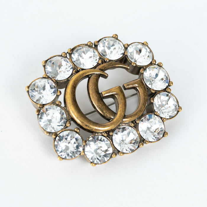 Gucci aged gold brooch with 'GG' logo and crystal stones on a white background